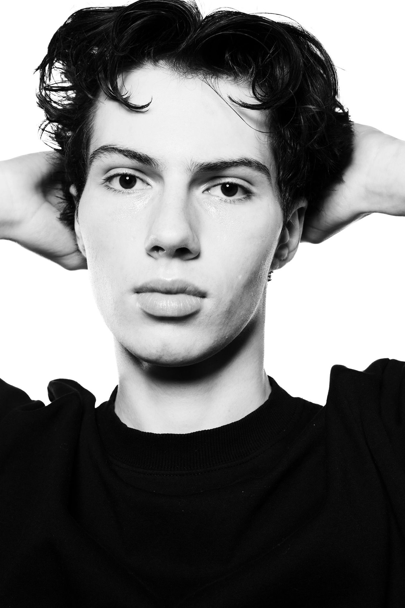 Monochromatic portrait of a young adult man with dark hair, hands behind his head, wearing a black shirt against a white backdrop.