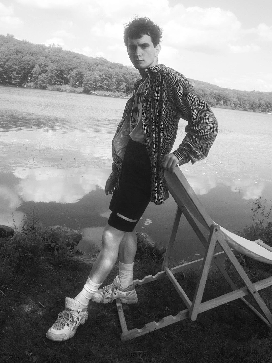 Black and white image of a young adult male in patterned shirt, shorts and sneakers, leaning against a wooden beach chair by a lake.