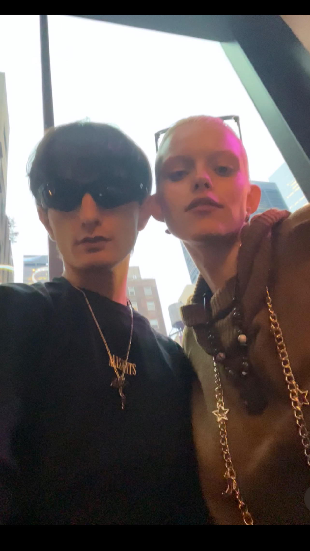 Two people pose for a selfie in front of a window with a city view. One wears sunglasses and a necklace, the other has short hair.