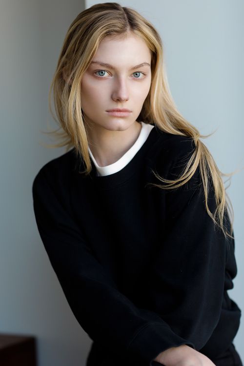 Blonde woman with blue eyes wearing a black sweatshirt with a white undershirt collar against a neutral studio background.
