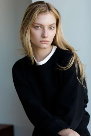 Blonde woman with blue eyes wearing a black sweatshirt with a white undershirt collar against a neutral studio background.