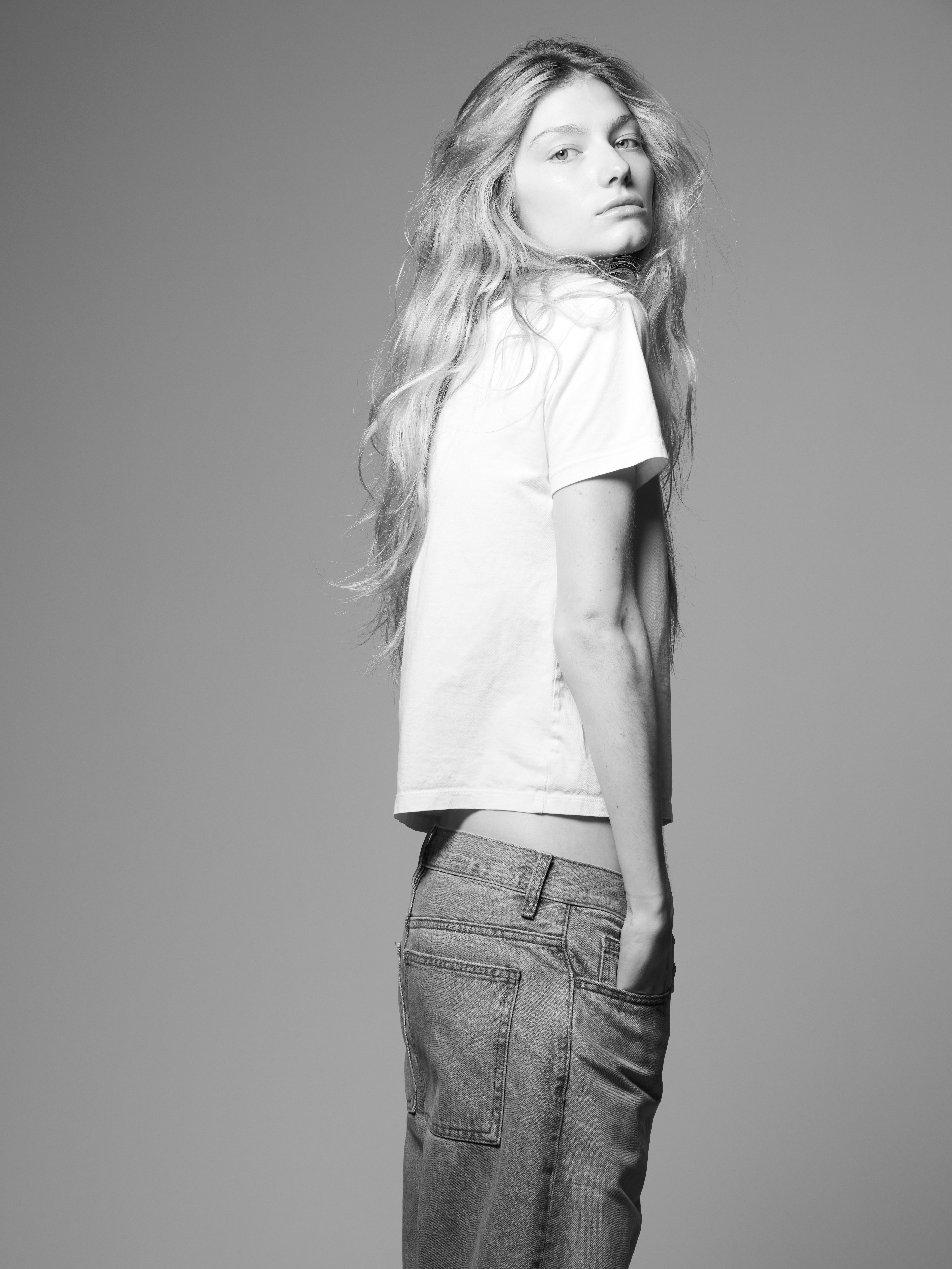 Black and white portrait of a woman with long wavy hair, wearing a white t-shirt and jeans, standing against a neutral grey background.
