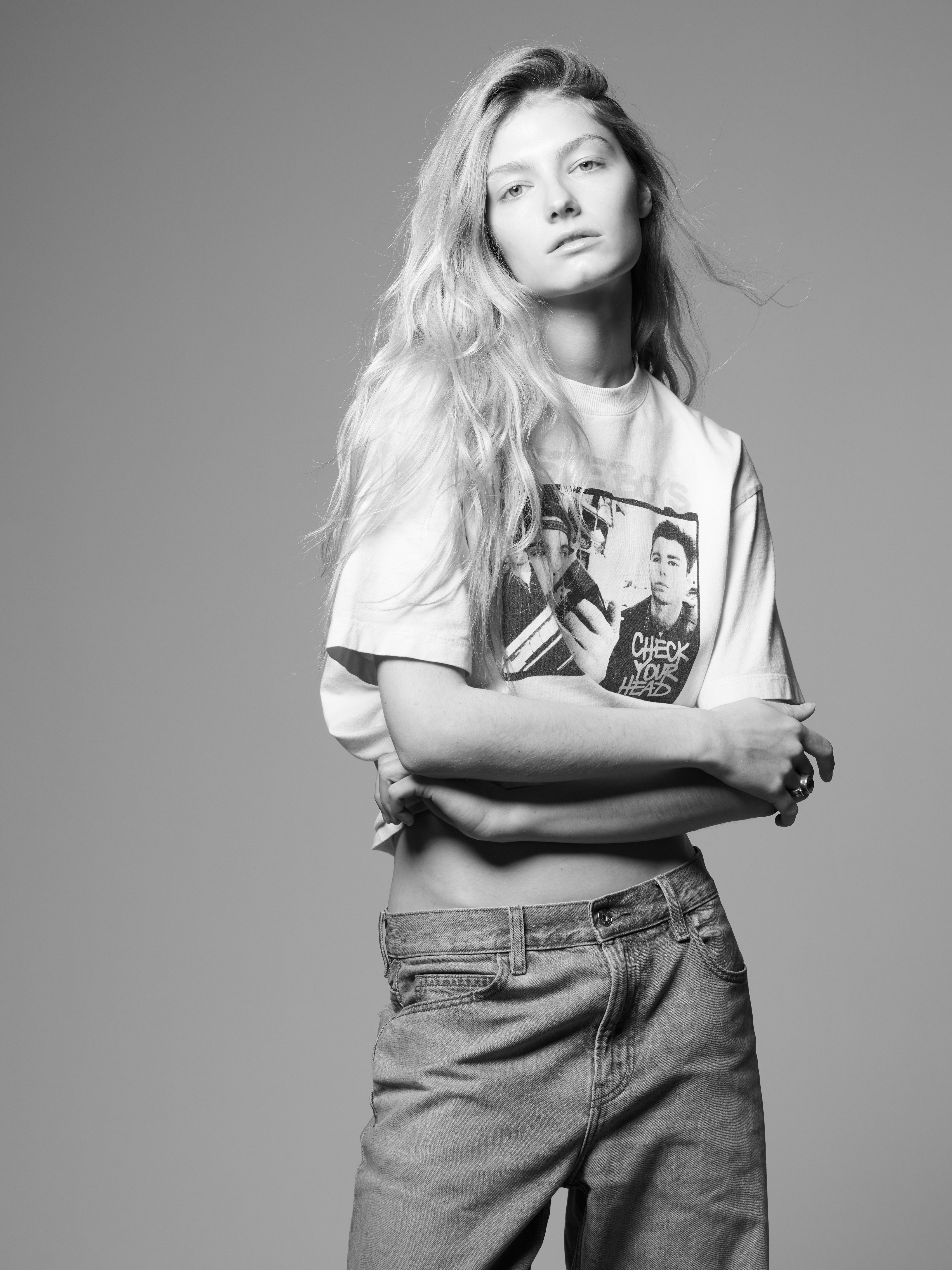 Black and white studio portrait of an adult woman with long blonde hair, wearing a graphic tee shirt and light-wash jeans, arms crossed.