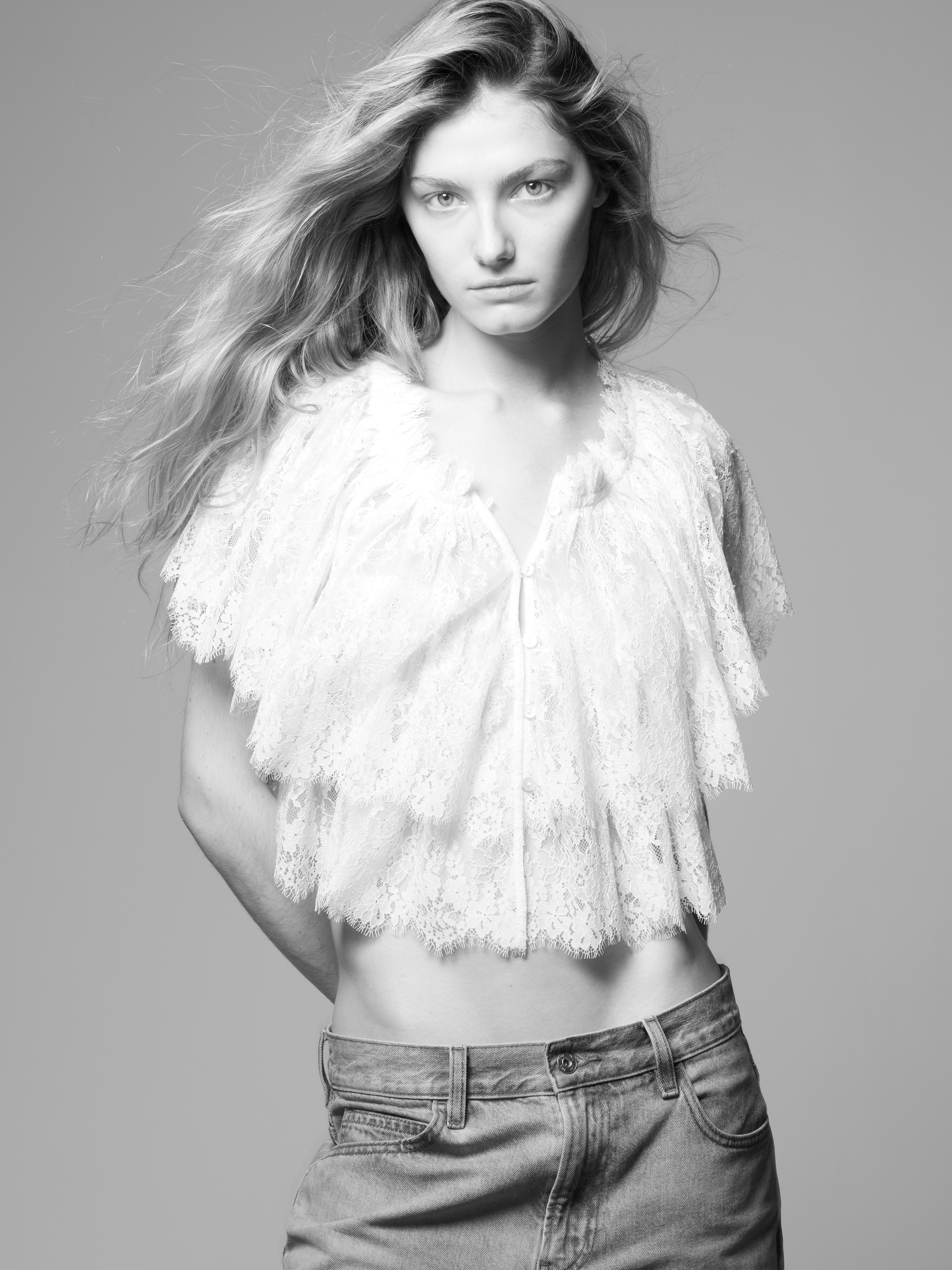 Monochrome fashion portrait of a young woman with flowing hair, wearing a white lace top and jeans against a grey background.
