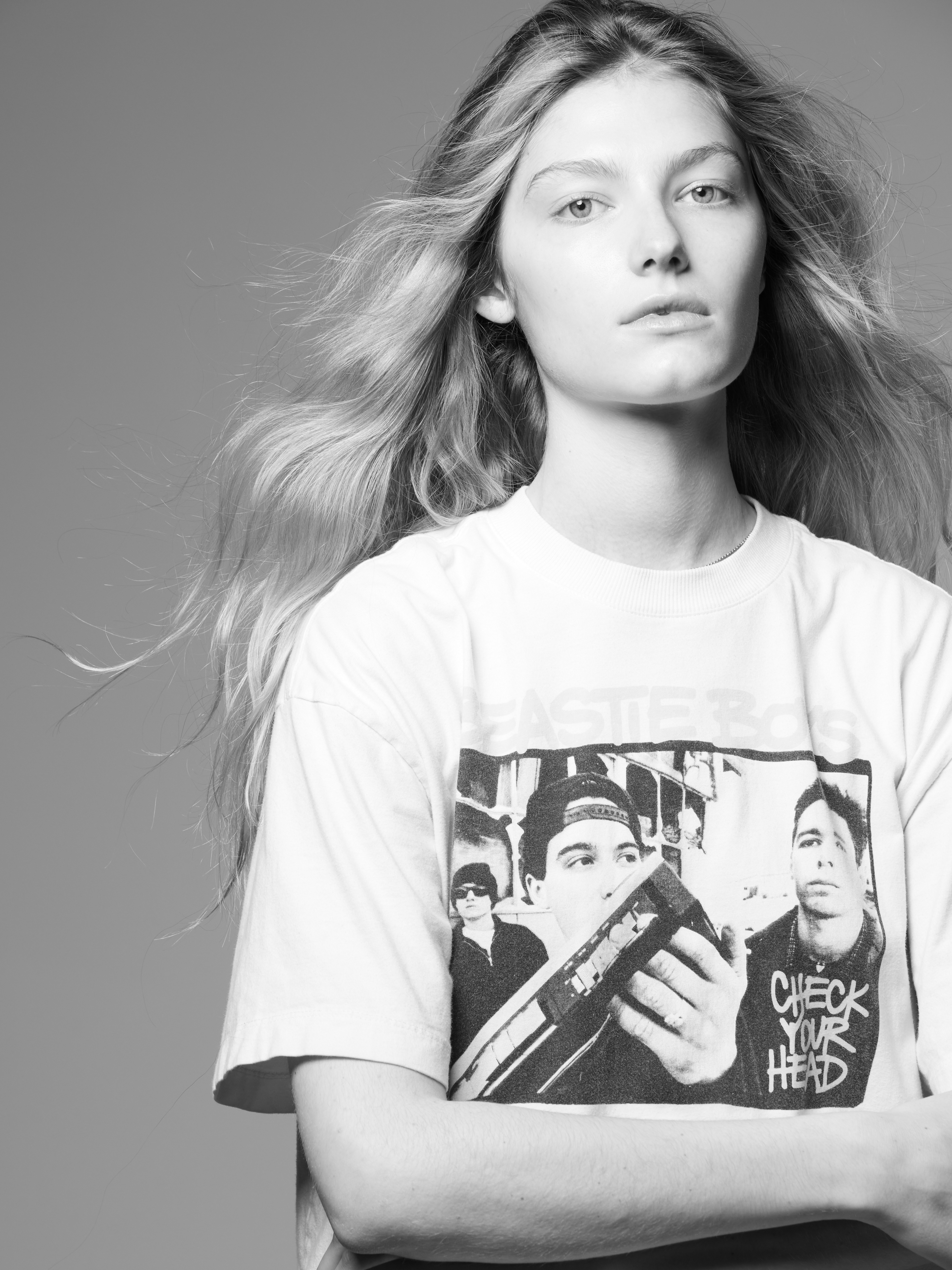 Black and white portrait of a young fair-skinned woman with long blonde hair, posing in a white graphic t-shirt against a gray background.