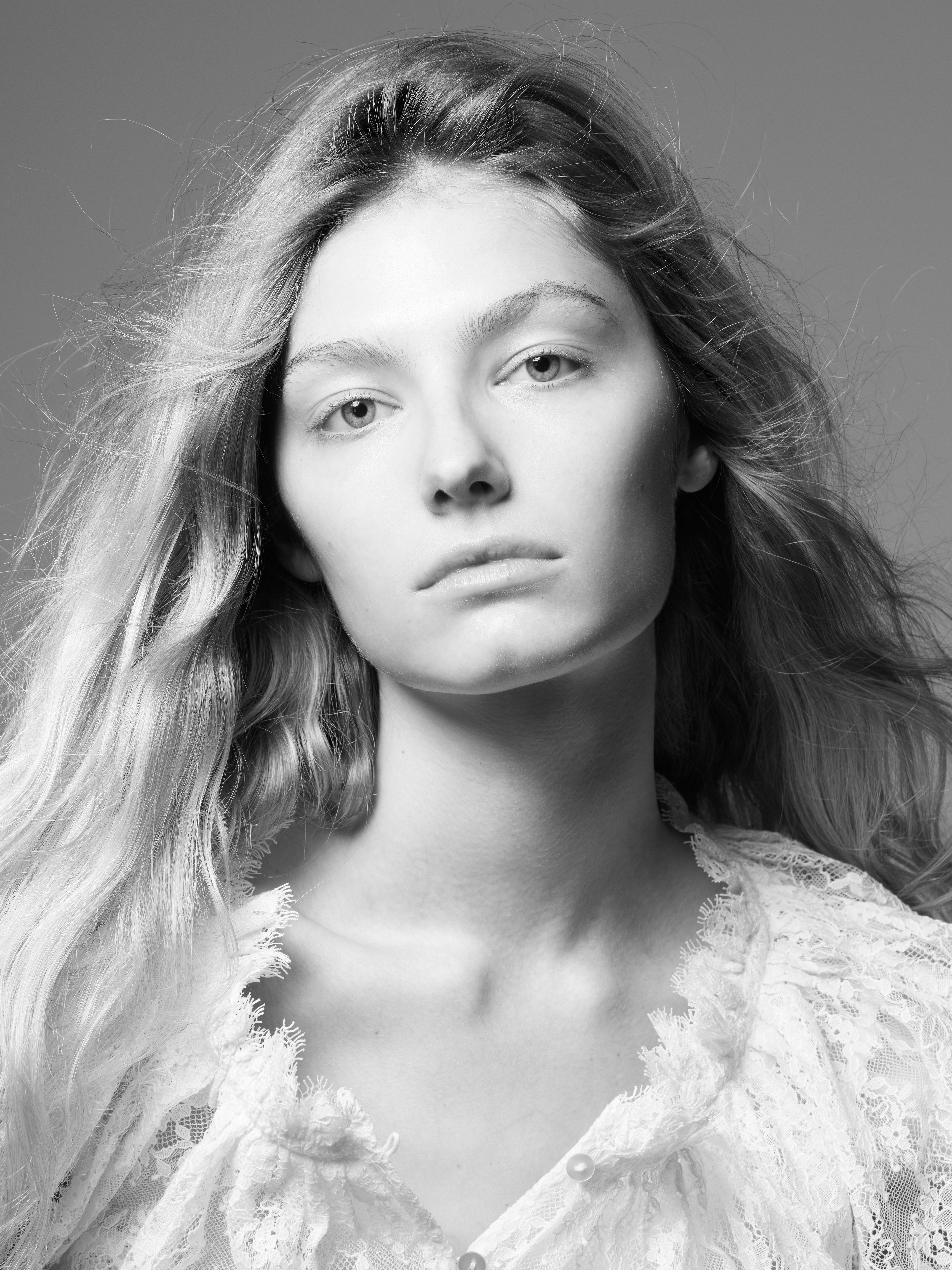 Black and white portrait of a young adult woman with long blonde wavy hair wearing a lace blouse against a grey studio background.