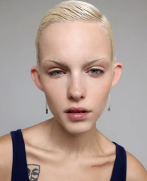 Close-up portrait of a model with short blonde hair and bleached eyebrows wearing safety pin earrings and a navy tank top.