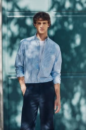 Young man with curly hair wearing a blue striped button-down shirt and dark navy trousers against a teal door in dappled sunlight.