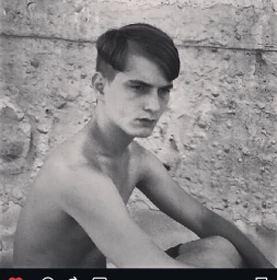 Monochrome portrait of a young man with styled dark hair seated against a textured concrete wall