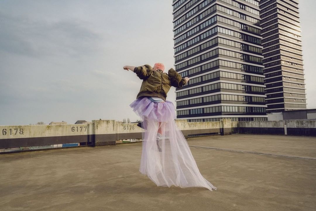 A person jumps in a purple tutu on a rooftop with apartment buildings in the background on an overcast day, wearing an olive jacket.