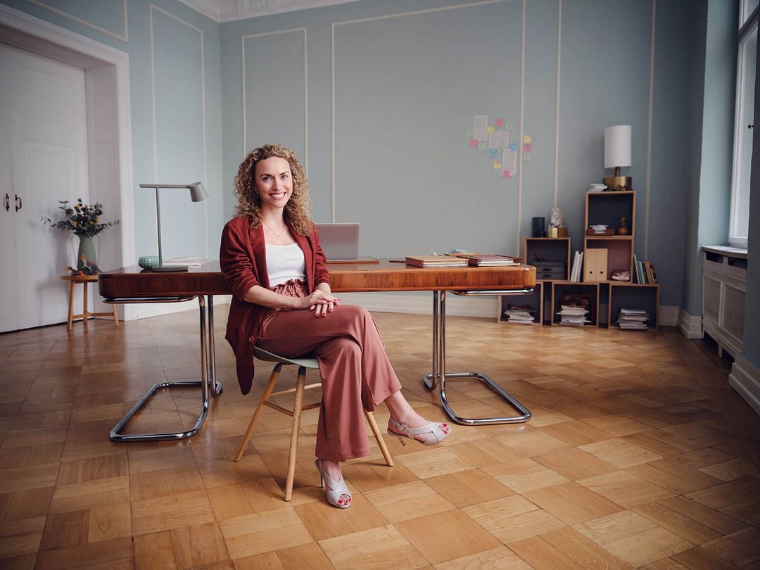 A woman sits cross-legged in rust-colored pants and a matching jacket, smiling in an office setting with a wooden desk and blue walls.
