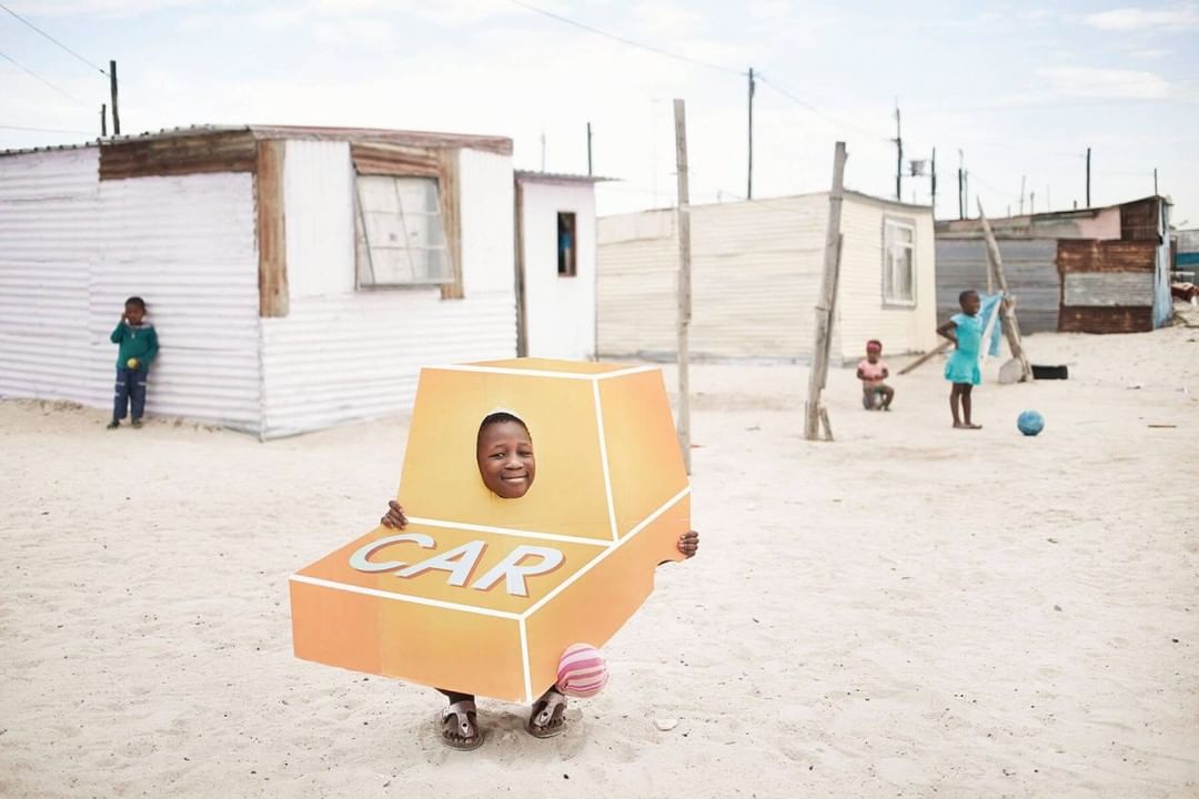 A smiling child stands in a dusty neighborhood wearing a cutout cardboard car costume, with other children and shacks in the background.