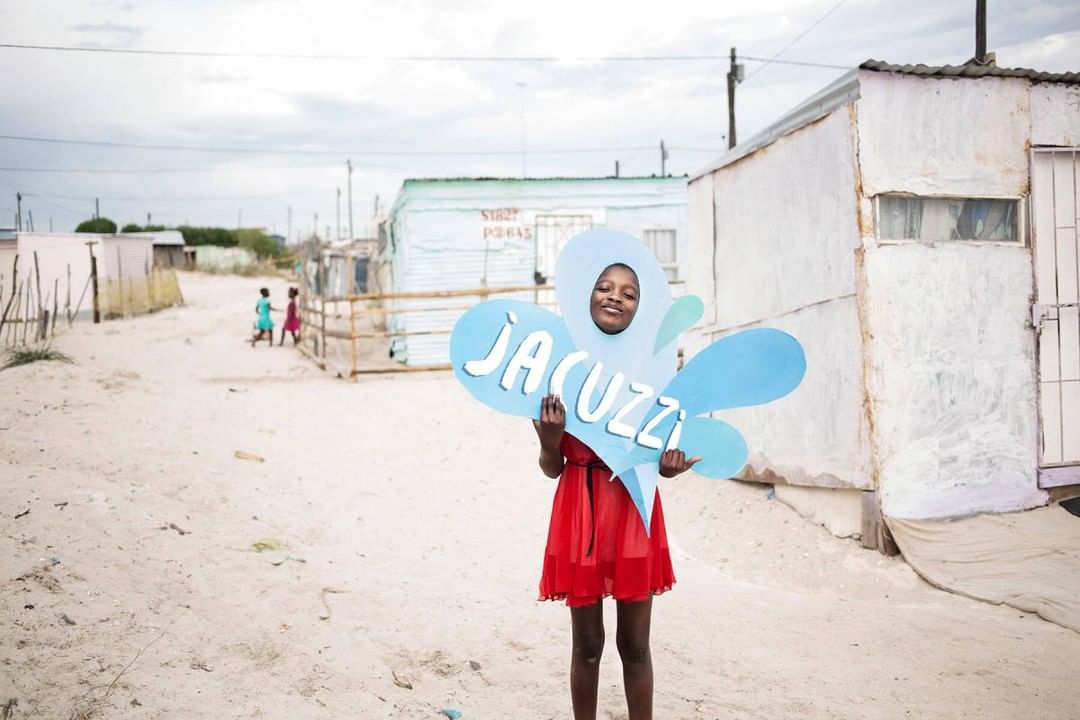 Young girl stands in a rural setting, holding a large blue 'Jacuzzi' sign over her head, creating a whimsical portrait with a hopeful expression.