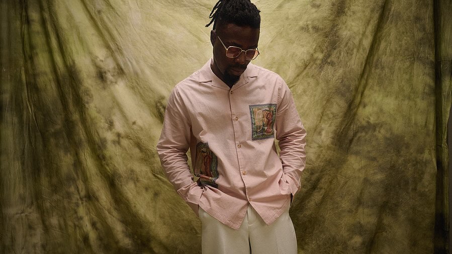 An adult male in a light pink shirt with artwork detail, and white pants, stands in front of a mottled green background with his hands in his pockets.
