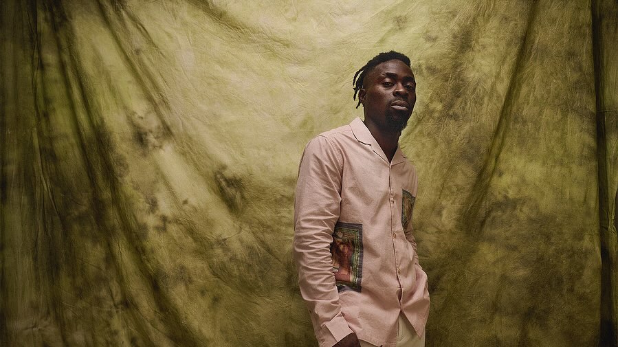Portrait of a Black adult male in a light-pink patterned shirt against a mottled olive-green backdrop in a photo studio.