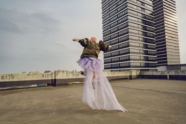 Person with pink hair jumping in a puffy olive green jacket and a long sheer lavender tulle skirt on a concrete rooftop.