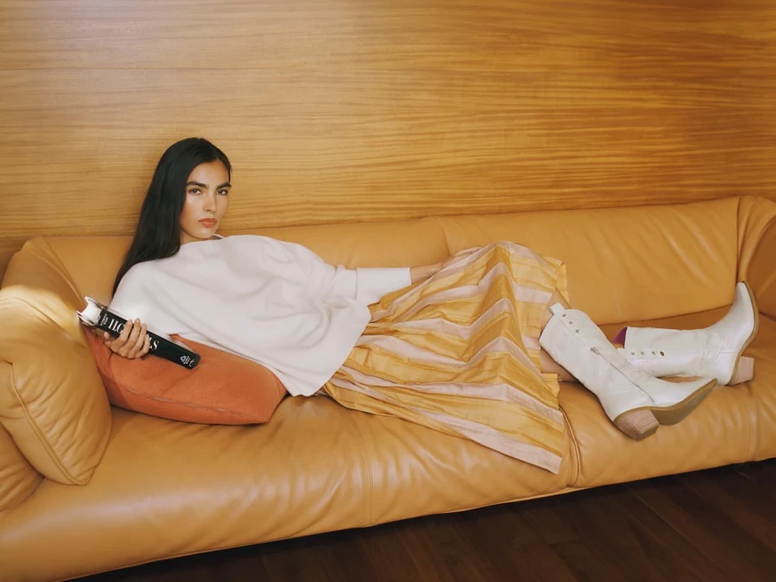 Adult woman reclines on an orange couch in a light-filled room wearing a white top, striped skirt, and white boots, and holding a black book.