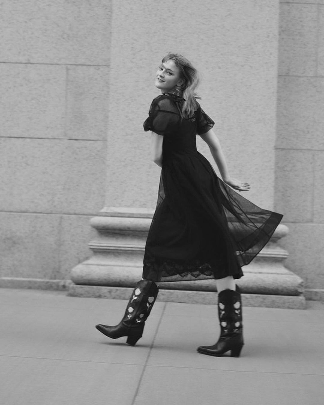 Monochrome portrait of a young woman in a dark dress and decorative cowboy boots walking near a stone wall and smiling back at the camera.