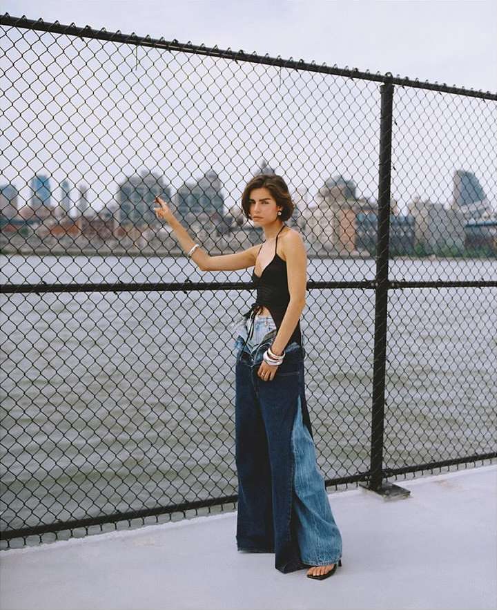 Woman poses in wide-leg jeans with city skyline backdrop. A chain-link fence is in the foreground, and the model looks toward the left.