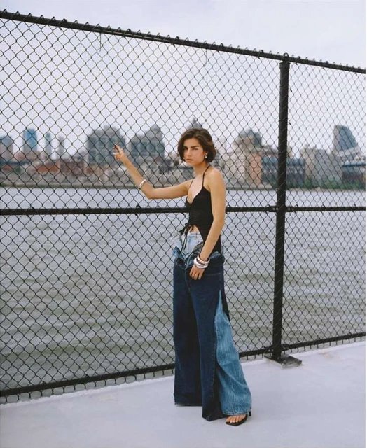 Woman poses in wide-leg jeans with city skyline backdrop. A chain-link fence is in the foreground, and the model looks toward the left.