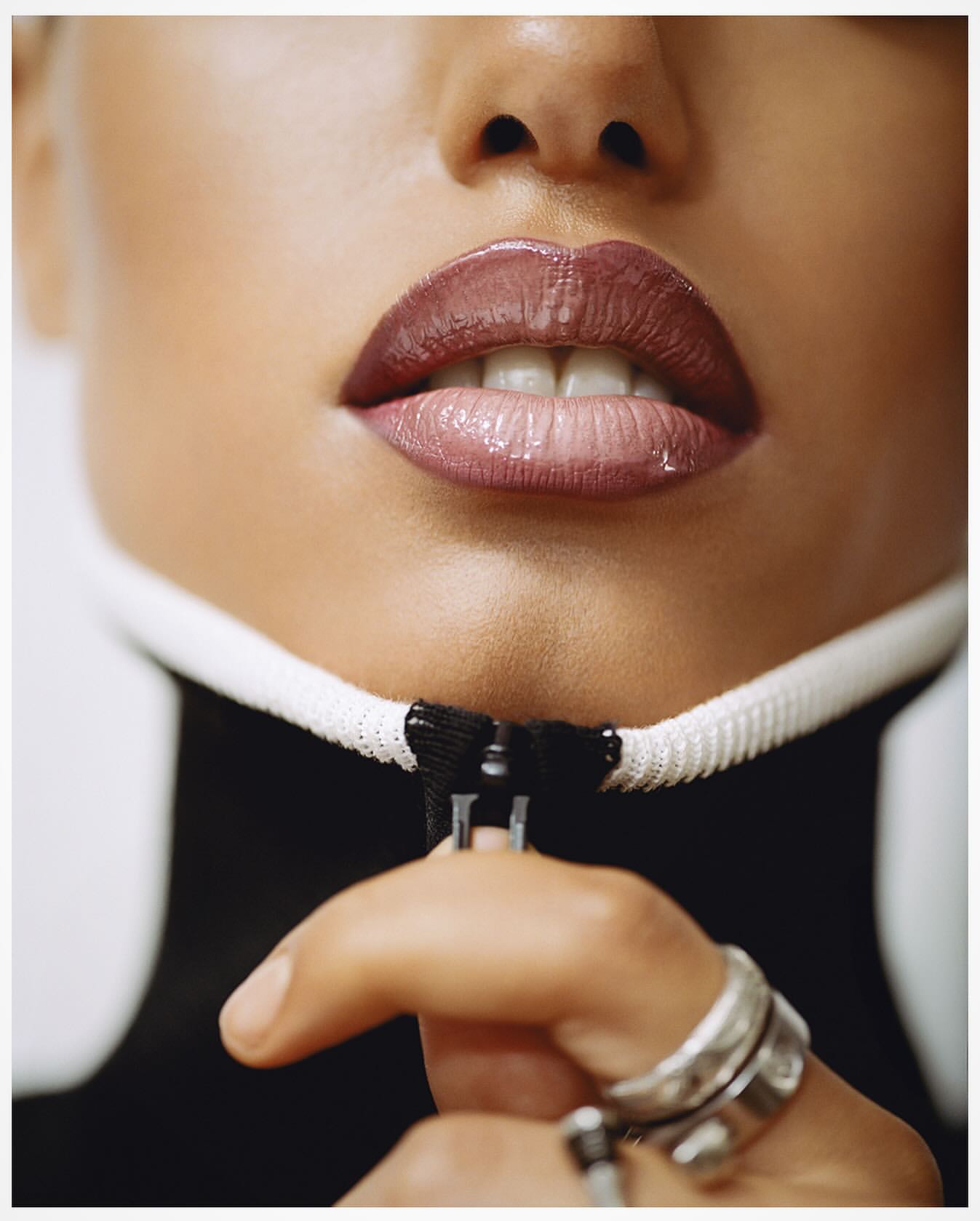 Close-up studio shot of an adult woman with hand on the zip of a black and white top, displaying glossy lip makeup and rings.