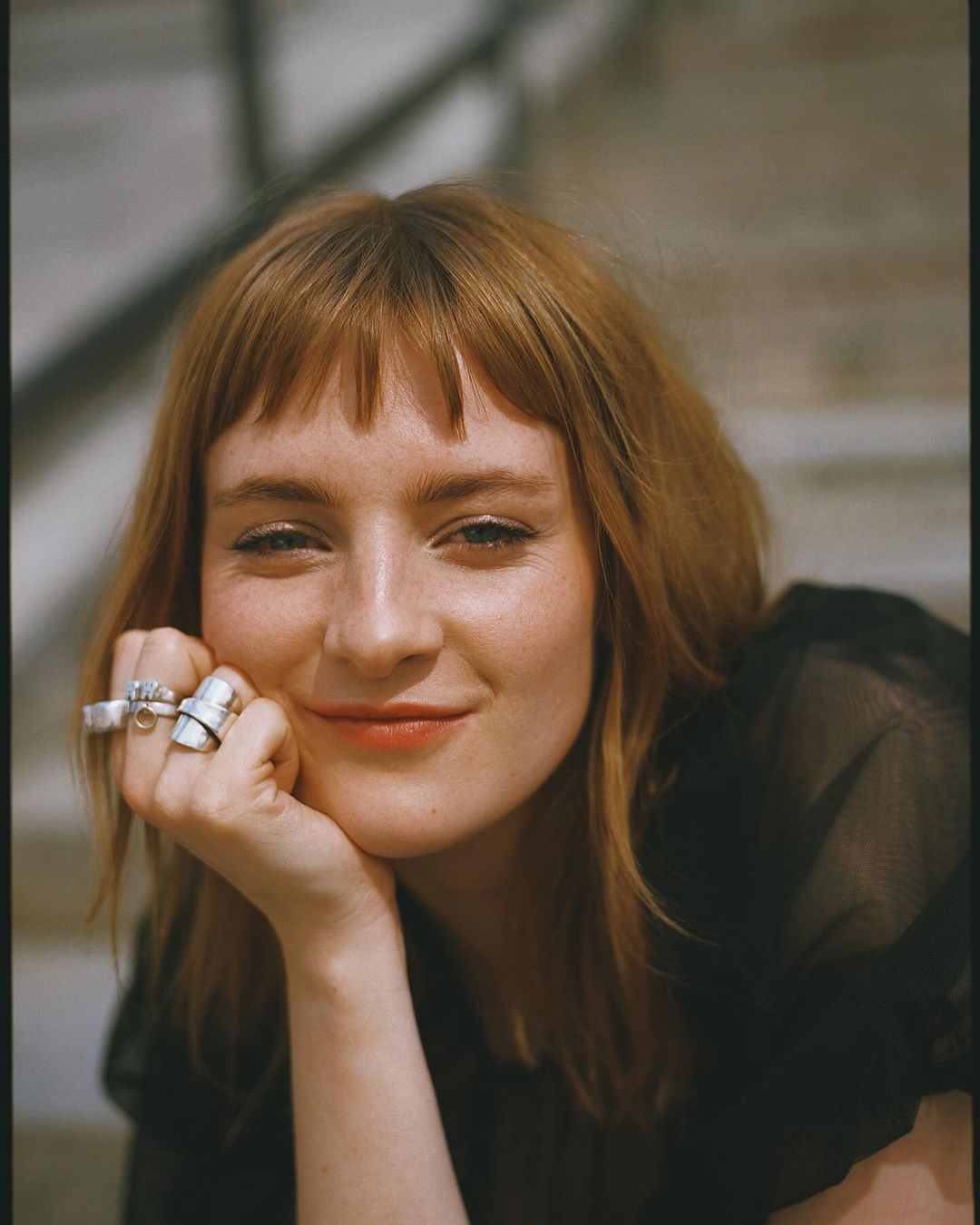 Portrait of a smiling young woman with auburn hair, wearing a sheer black top, posing with her hand on her face outside.