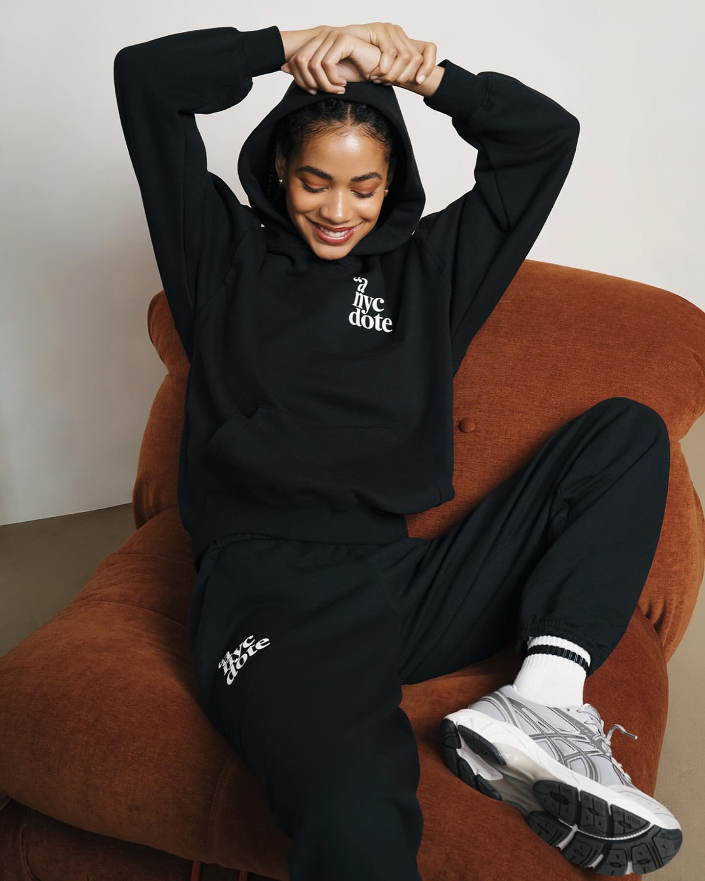 A Black woman sits in an indoor studio setting, wearing a black hoodie and sweatpants, with sneakers. The scene has neutral tones.