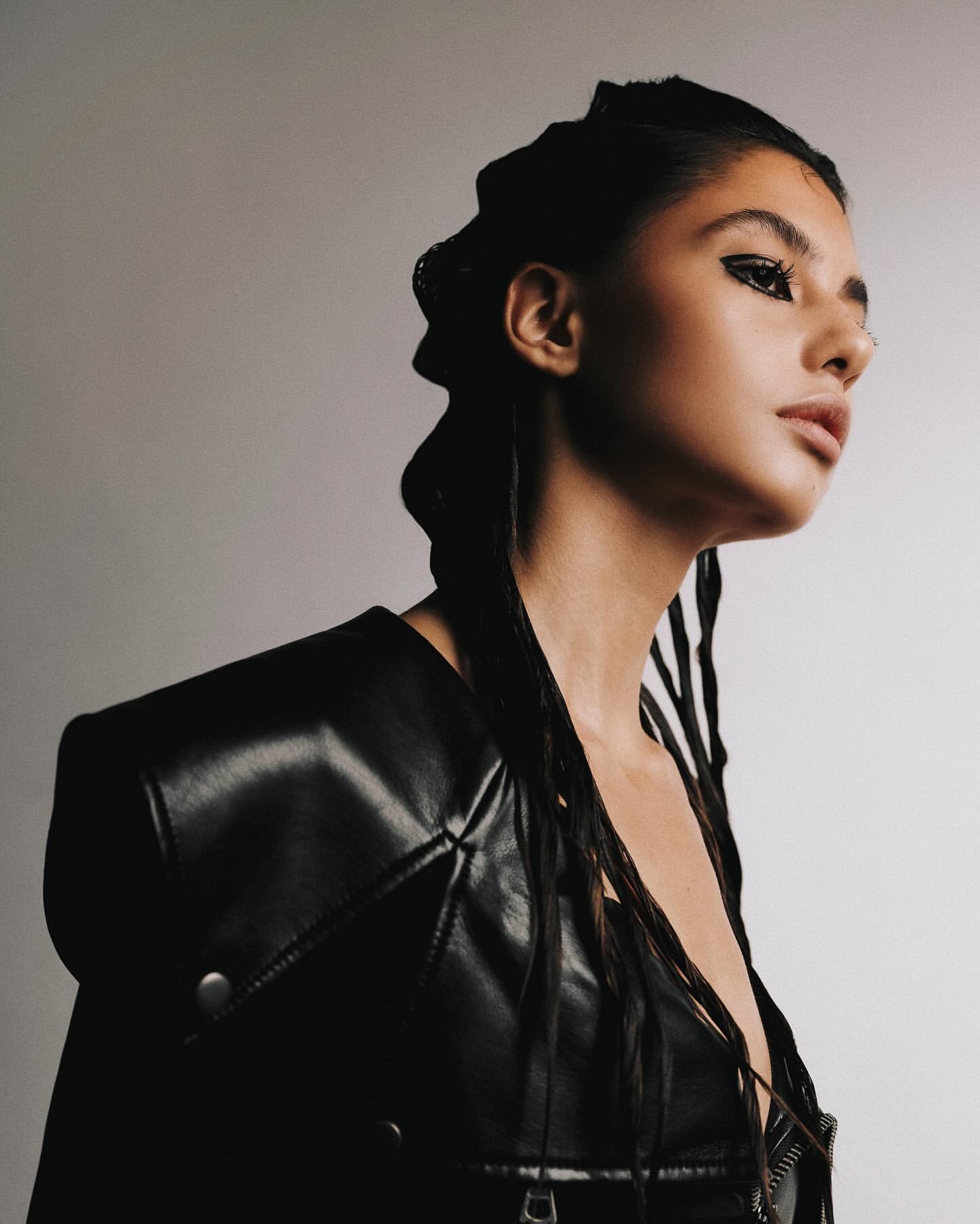 Portrait of a young woman in a black leather jacket with edgy eye makeup and braided hairstyle, posing in a studio setting on white background.
