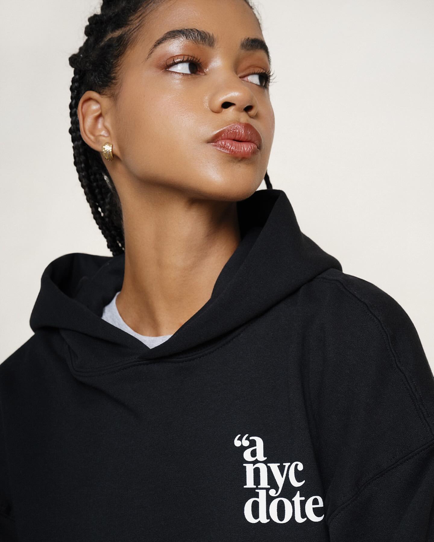 A young woman poses wearing a black hoodie in a studio setting with neutral background, showcasing beauty makeup and casual fashion.