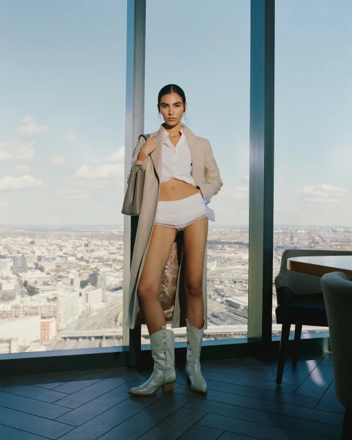 A woman in a tan coat, white shirt, and underwear stands in front of a tall building window overlooking a city.