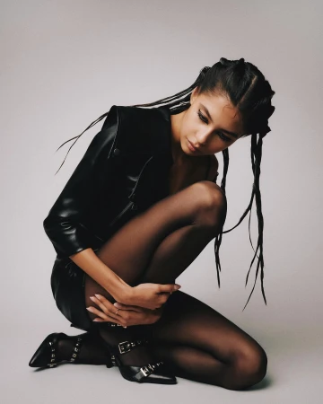 Young woman in a black leather jacket and skirt kneeling, wearing black tights and buckled heels against a neutral backdrop in a studio shot.