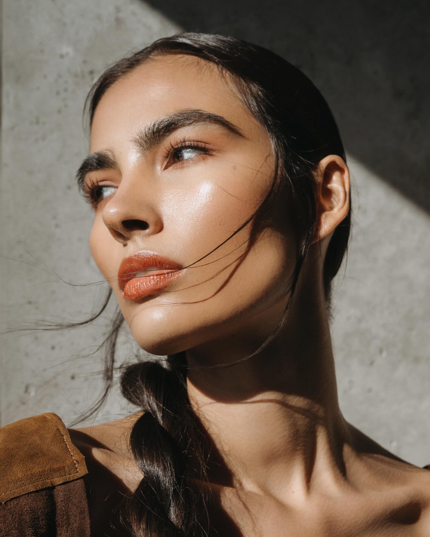 Portrait of a brunette woman with sleek hair in a braid, wearing a brown top, and posing in a room with harsh sunlight and shadows.