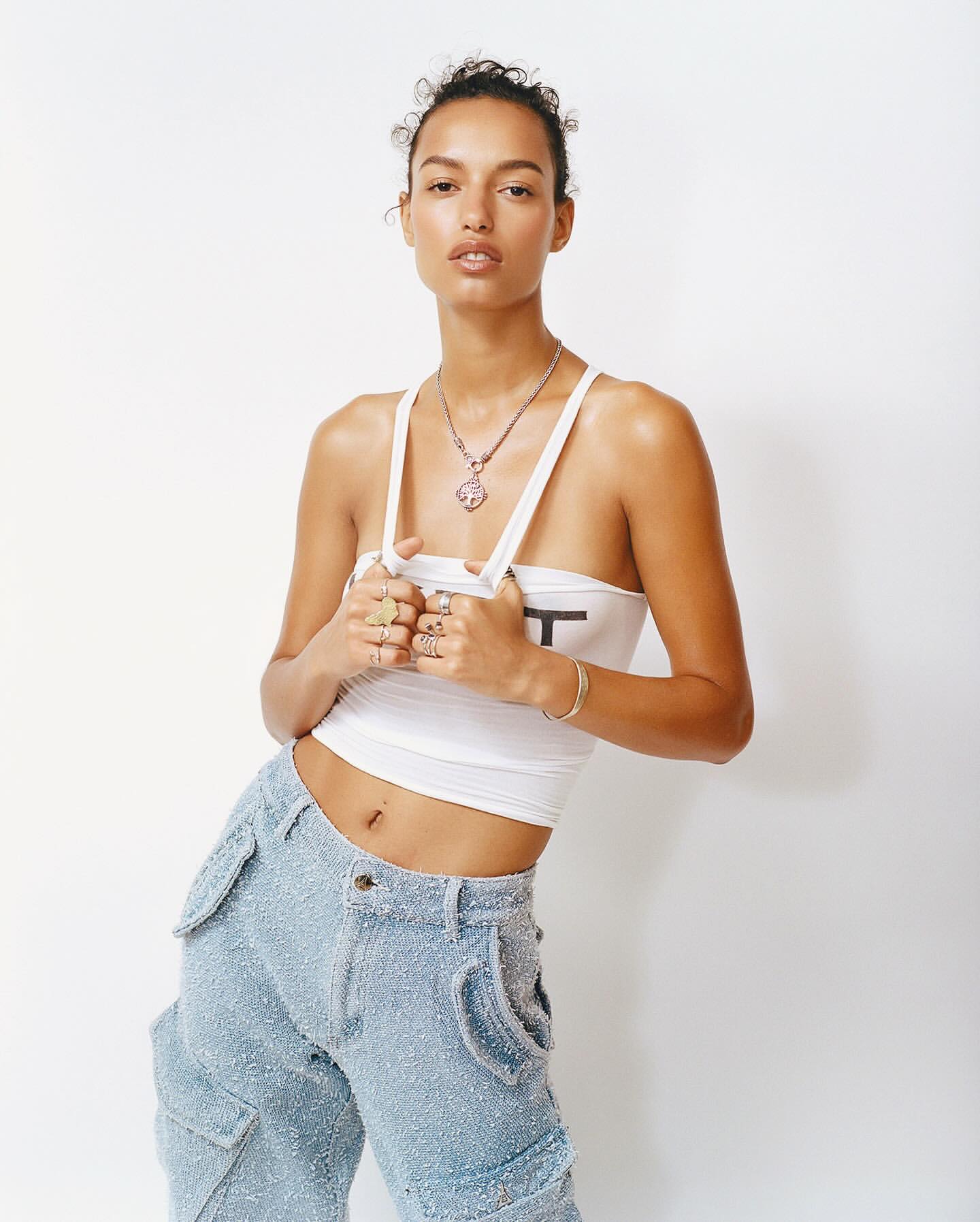 A slender woman poses in a white top and blue cargo pants against a white backdrop, accentuating her stylish, fashionable look.