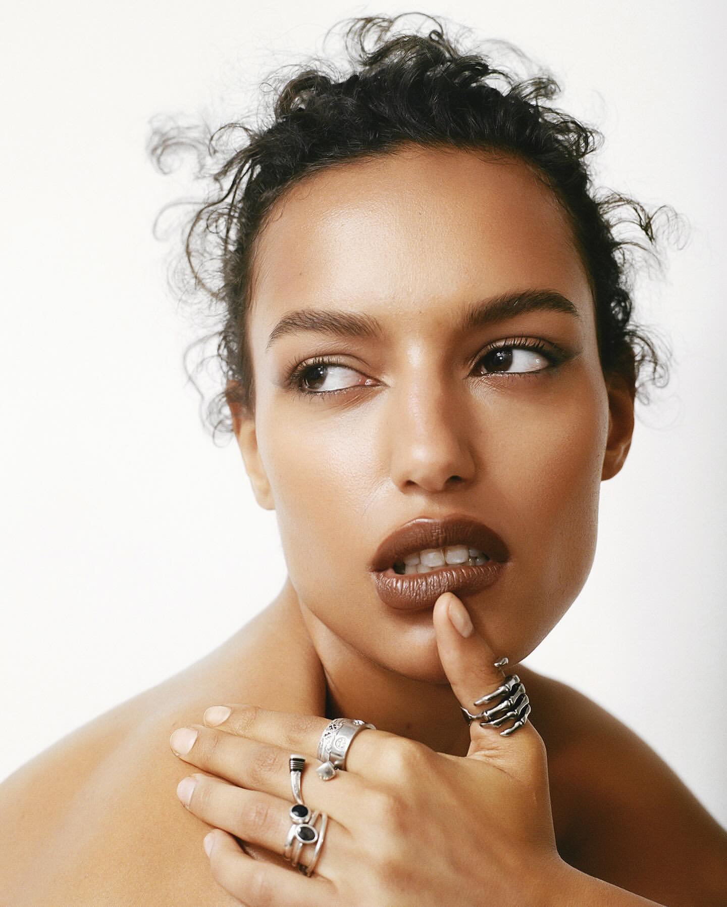 Close-up portrait of an adult woman with dark curly hair and a variety of silver rings on her fingers, set against a clean white backdrop.