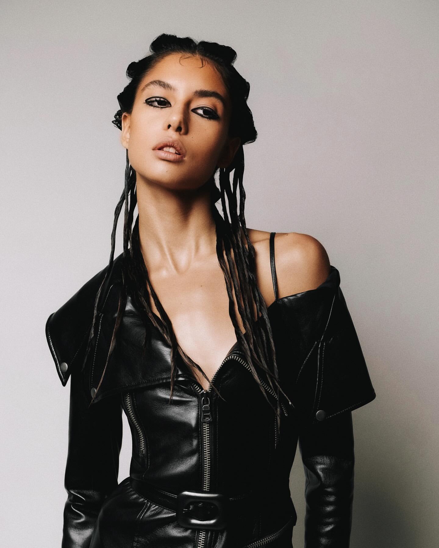 Portrait of a young adult woman with dark hair and dramatic eye makeup posing in a black leather jacket against a light background.