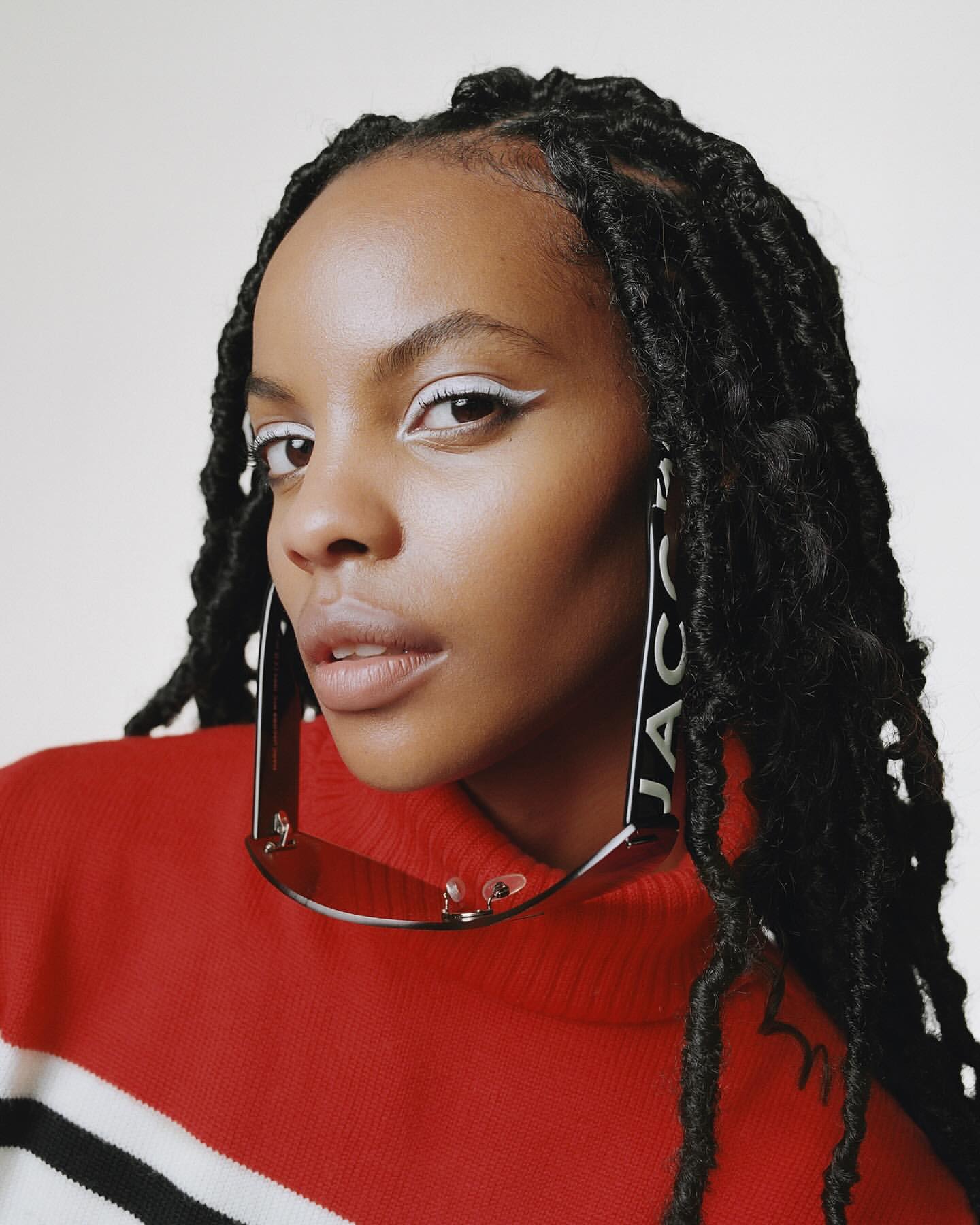 Close-up shot of a woman with dark braided hair, wearing a red and white sweater and sunglasses, in a brightly lit, neutral studio.