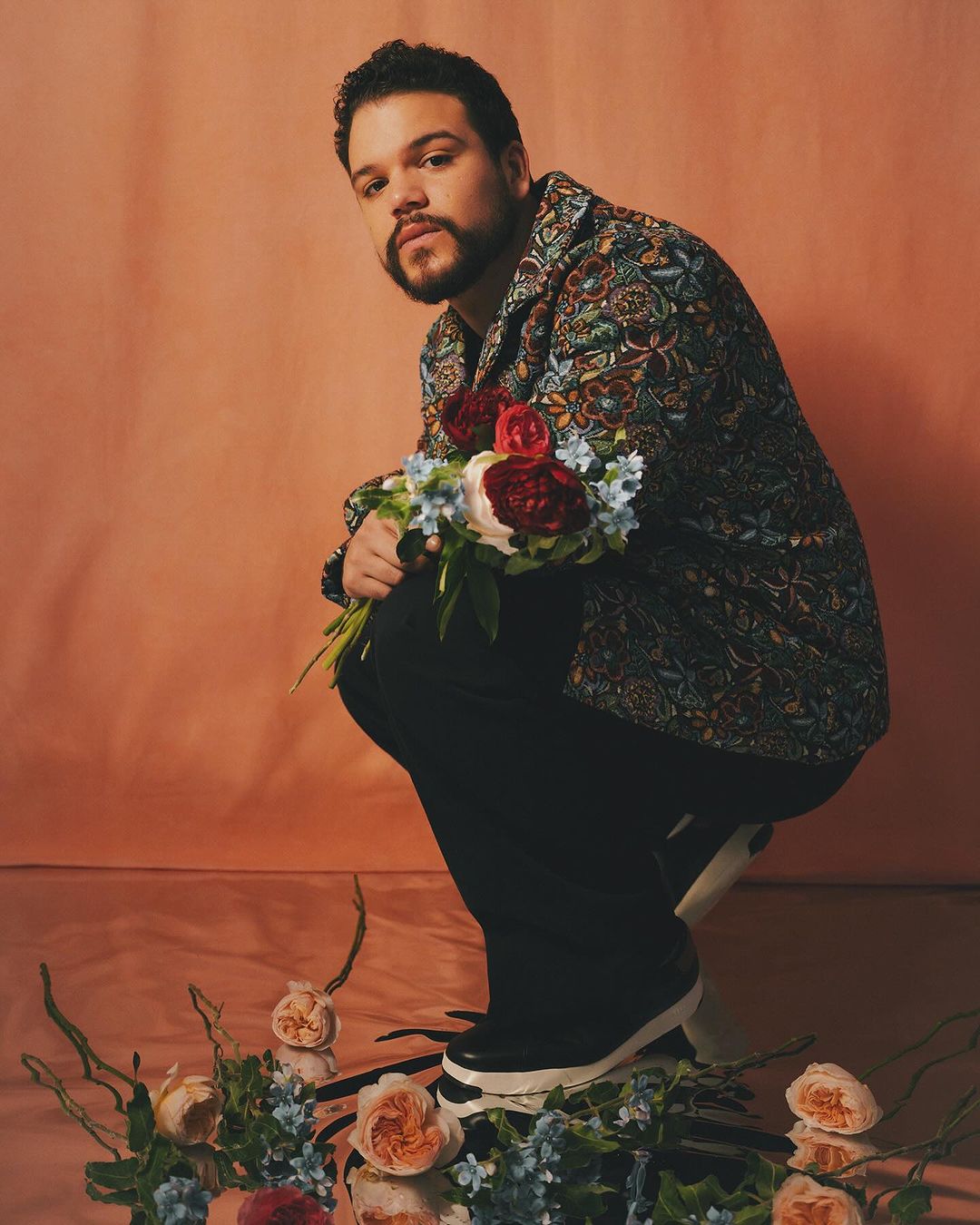 Man squats in a patterned jacket with flowers in a photo studio setting. Roses and other flowers scatter across the reflective floor.