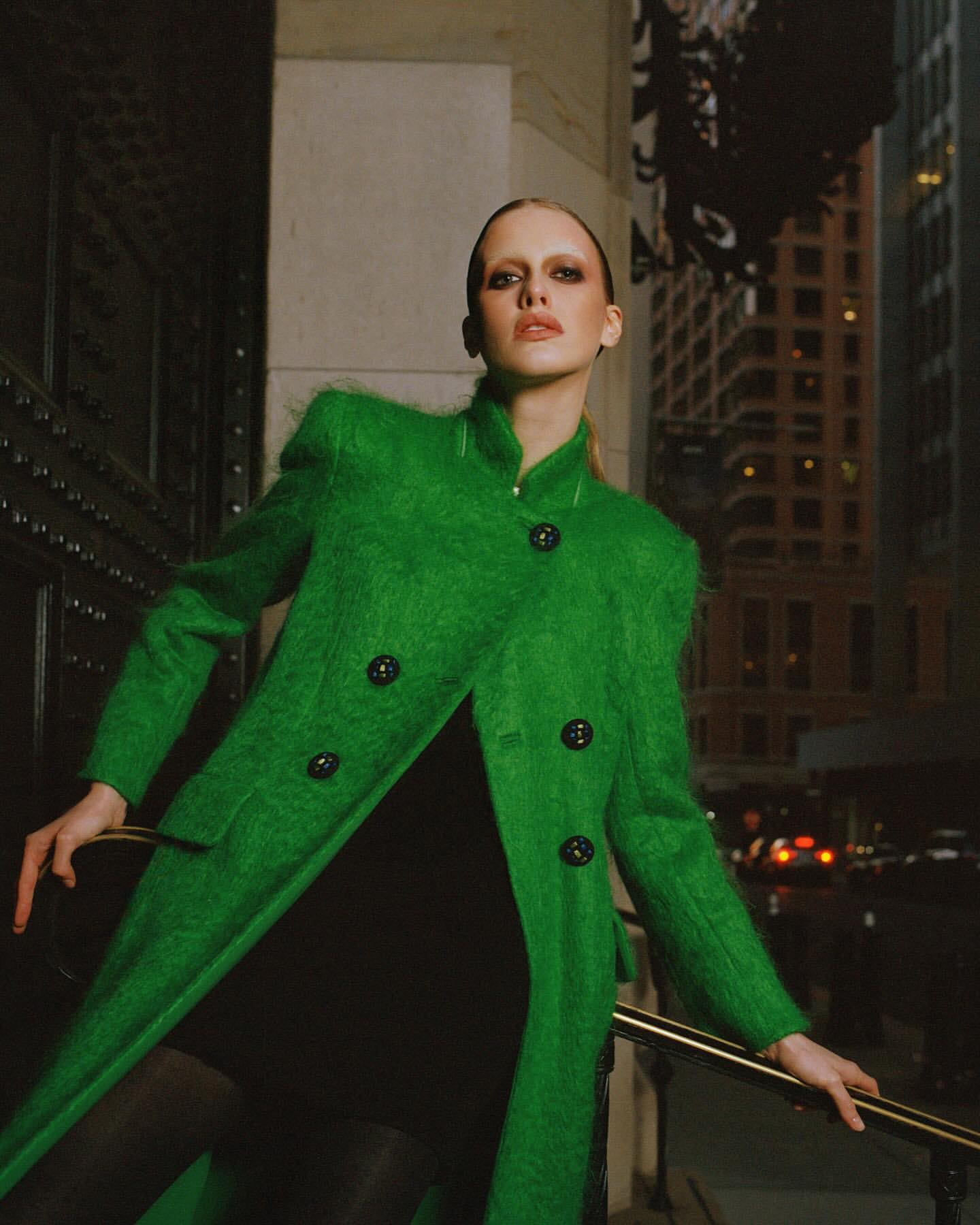 A stylish woman in a vibrant green coat poses against an urban backdrop, exuding confidence and sophistication in an eye-level fashion shot.