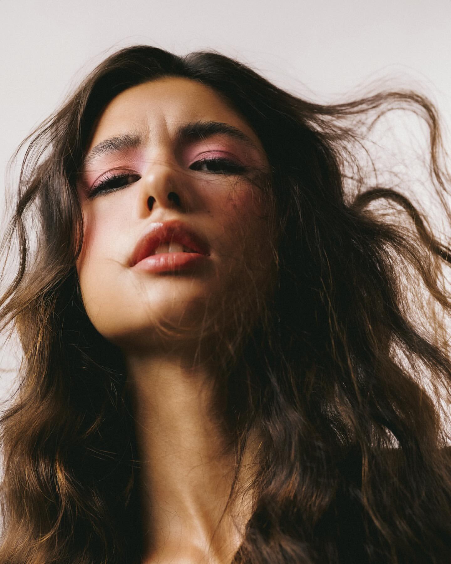Portrait of a woman with wavy brown hair and pink eye shadow, looking directly at the camera from a low angle against a white background.