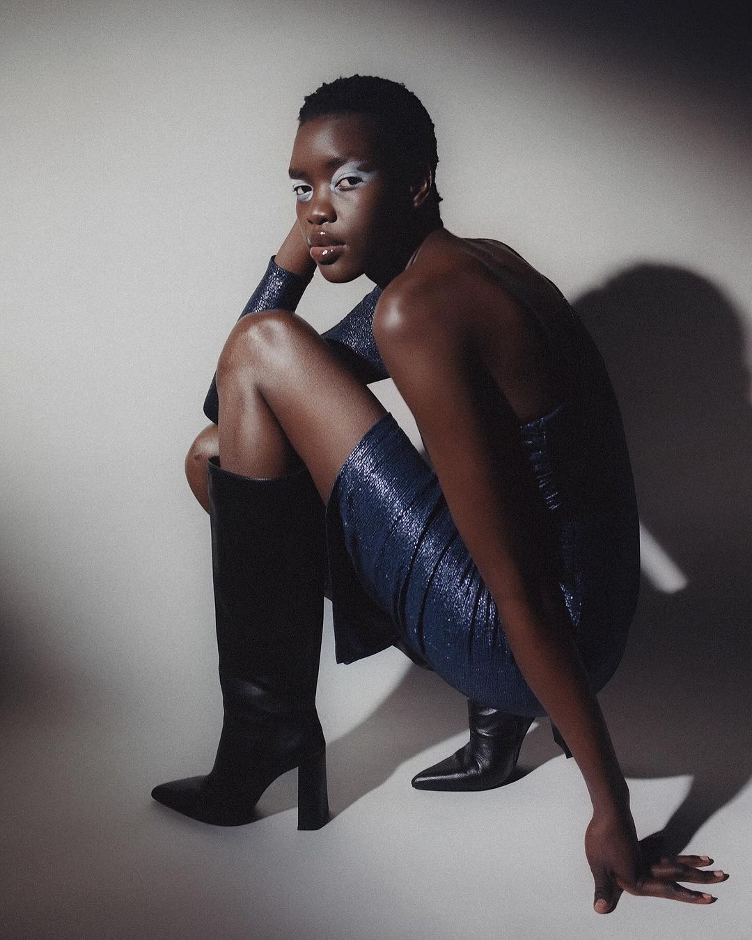 Dark-skinned model poses in a blue dress and black boots in a studio, showcasing fashion and beauty in a stylish, full-body shot.