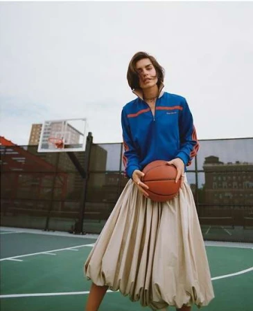 Female model poses with basketball on an outdoor court wearing a blue track jacket and large beige skirt against a city backdrop.