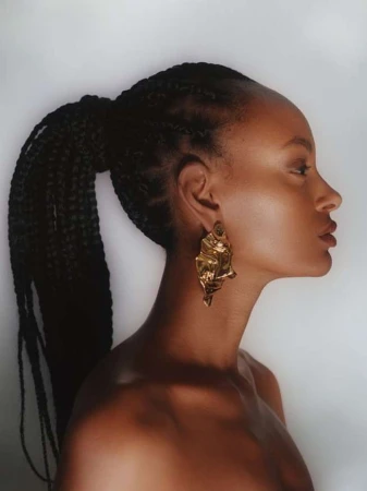 Profile portrait of an adult female with dark complexion and braided hair, wearing a sculptural gold earring against a soft white background.