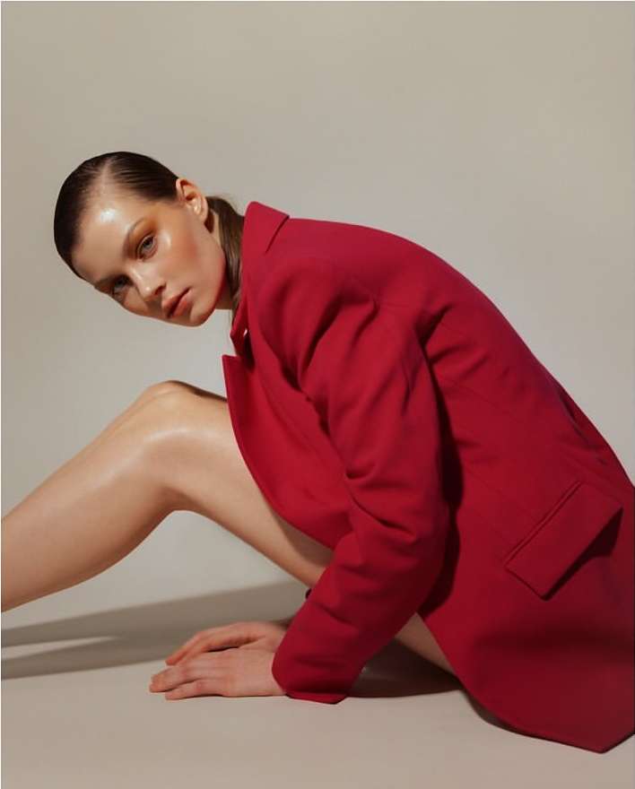 A fair-skinned adult woman in a red jacket poses indoors against a light background, exhibiting contemporary style with slicked-back hair.