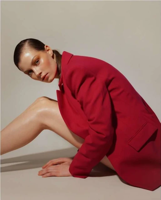 A fair-skinned adult woman in a red jacket poses indoors against a light background, exhibiting contemporary style with slicked-back hair.