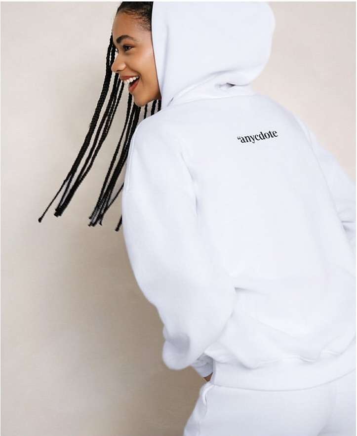 A woman smiles, turned to the side, wearing a white hoodie and sweatpants. The hoodie has black text on the upper back.