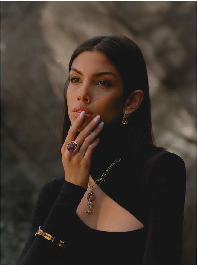 Woman in a black turtleneck poses with gemstone jewelry in an editorial fashion shot against a muted grey background.