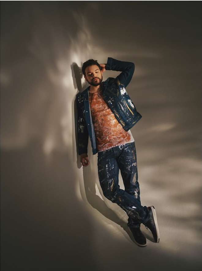 Portrait of a man in a denim jacket and paint-splattered jeans reclining against a neutral wall in a studio setting with soft lighting.