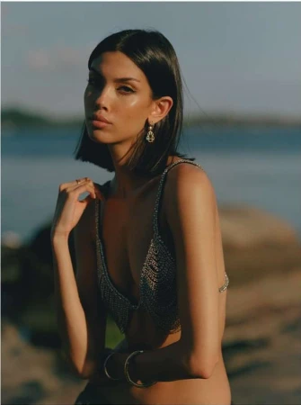 Close-up portrait of a dark-haired woman wearing a sparkly chainmail top and ornate earrings by water.