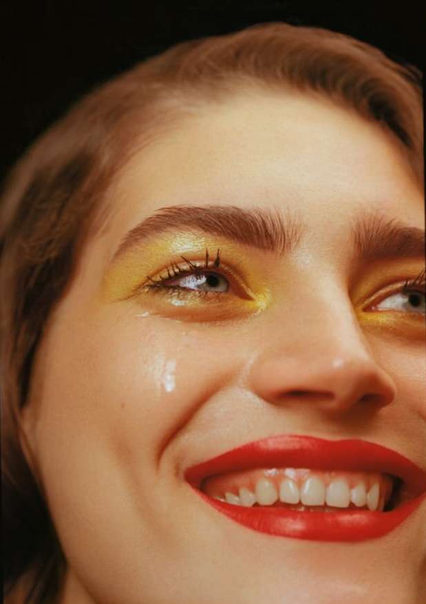 Close-up portrait of a woman with creative yellow eyeshadow, red lipstick, and a single tear, set against a dark, moody background.