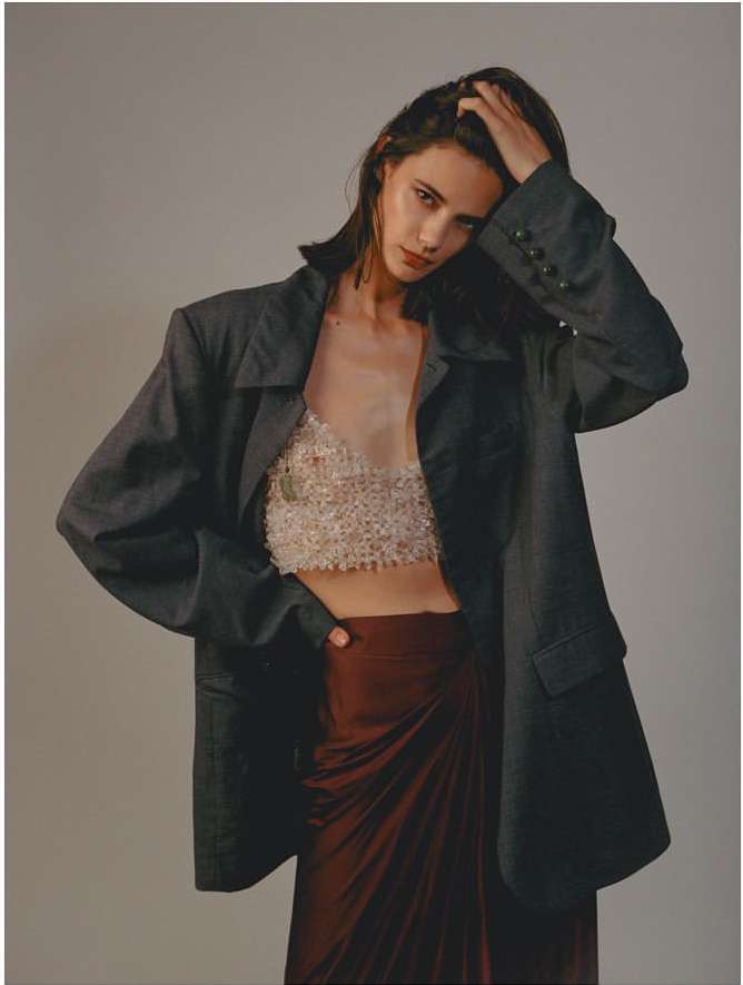 A woman in an oversized blazer poses in front of a neutral background, showcasing a textured top and maroon-colored skirt.